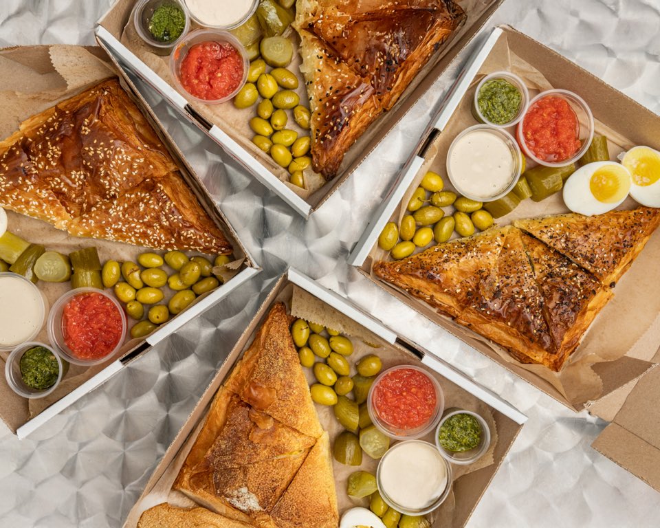 Four open BUBA party boxes arranged in a pinwheel, each with a large bureka, sauces, olives, pickles, and eggs.