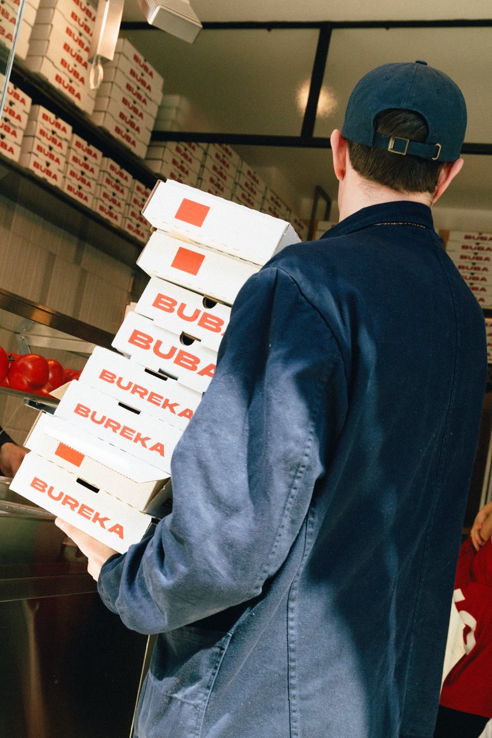 A staff member in navy workwear carries a tall stack of red-and-white BUBA boxes through the shop, shelves of boxes stacked behind.