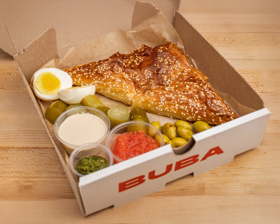 A sesame-crusted cheese bureka in a BUBA takeout box with a jammy egg, pickles, olives, tahini, crushed tomato, and schug.