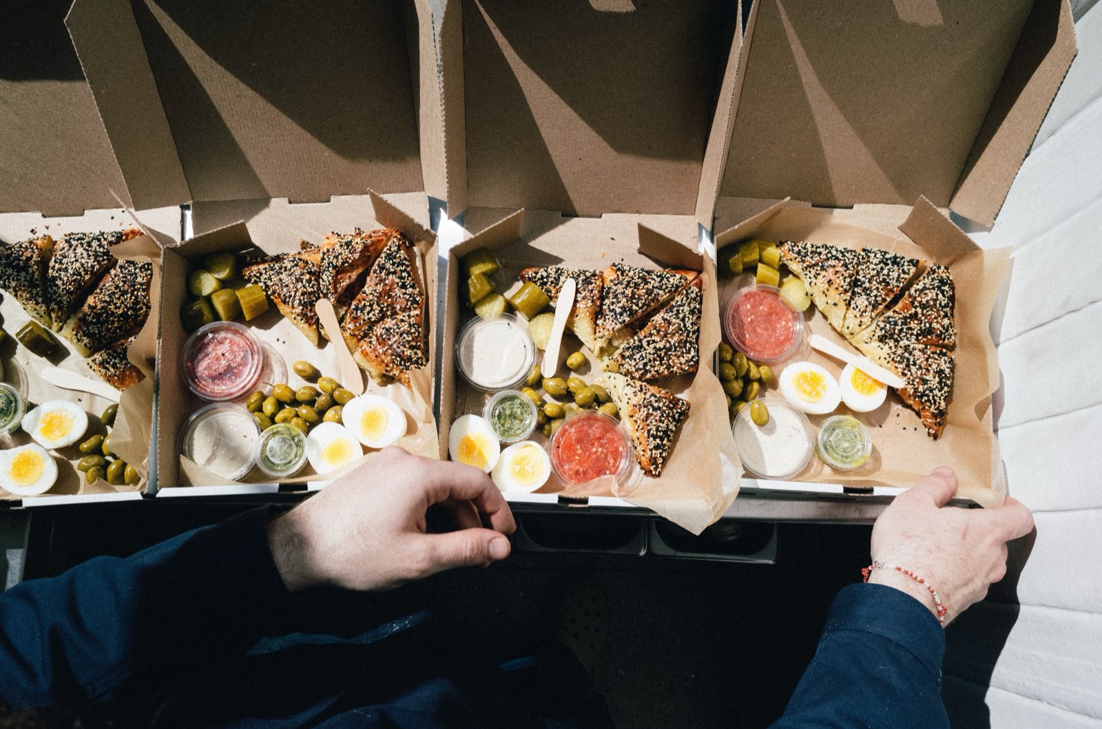 Four open BUBA boxes in bright sunlight, hands arranging burekas beside eggs, olives, pickles, and sauces.